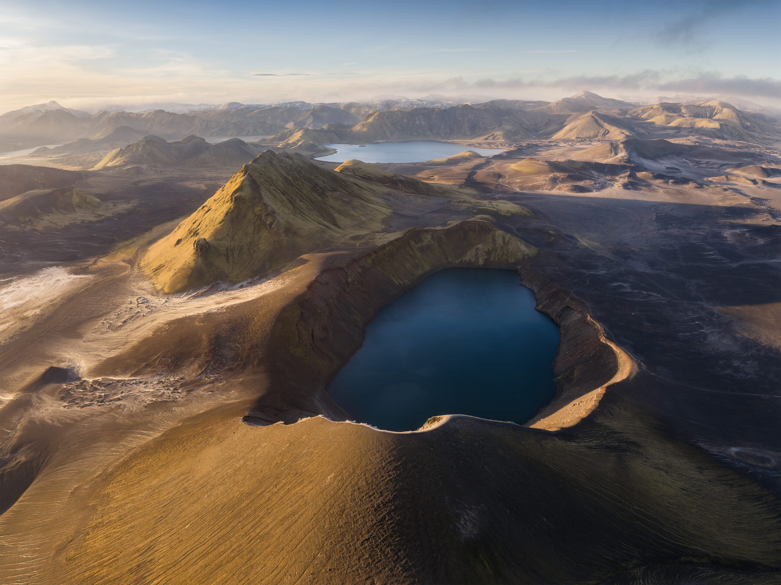 Veduta aerea di un lago di cratere blu scuro circondato da creste vulcaniche, con montagne e un altro lago sullo sfondo.
