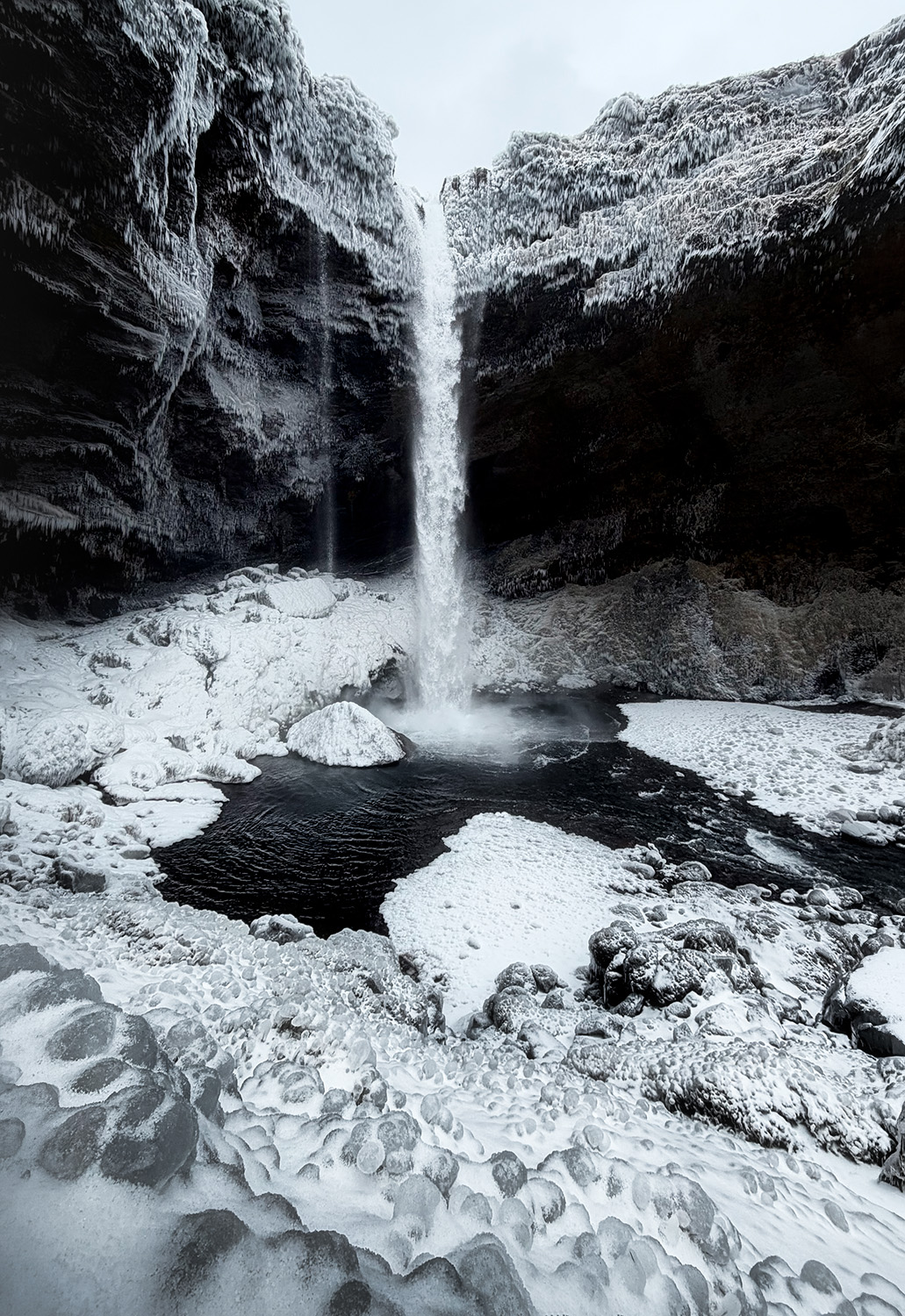 Cascata in una gola rocciosa coperta di neve e ghiaccio, con un piccolo specchio d'acqua scura in basso.