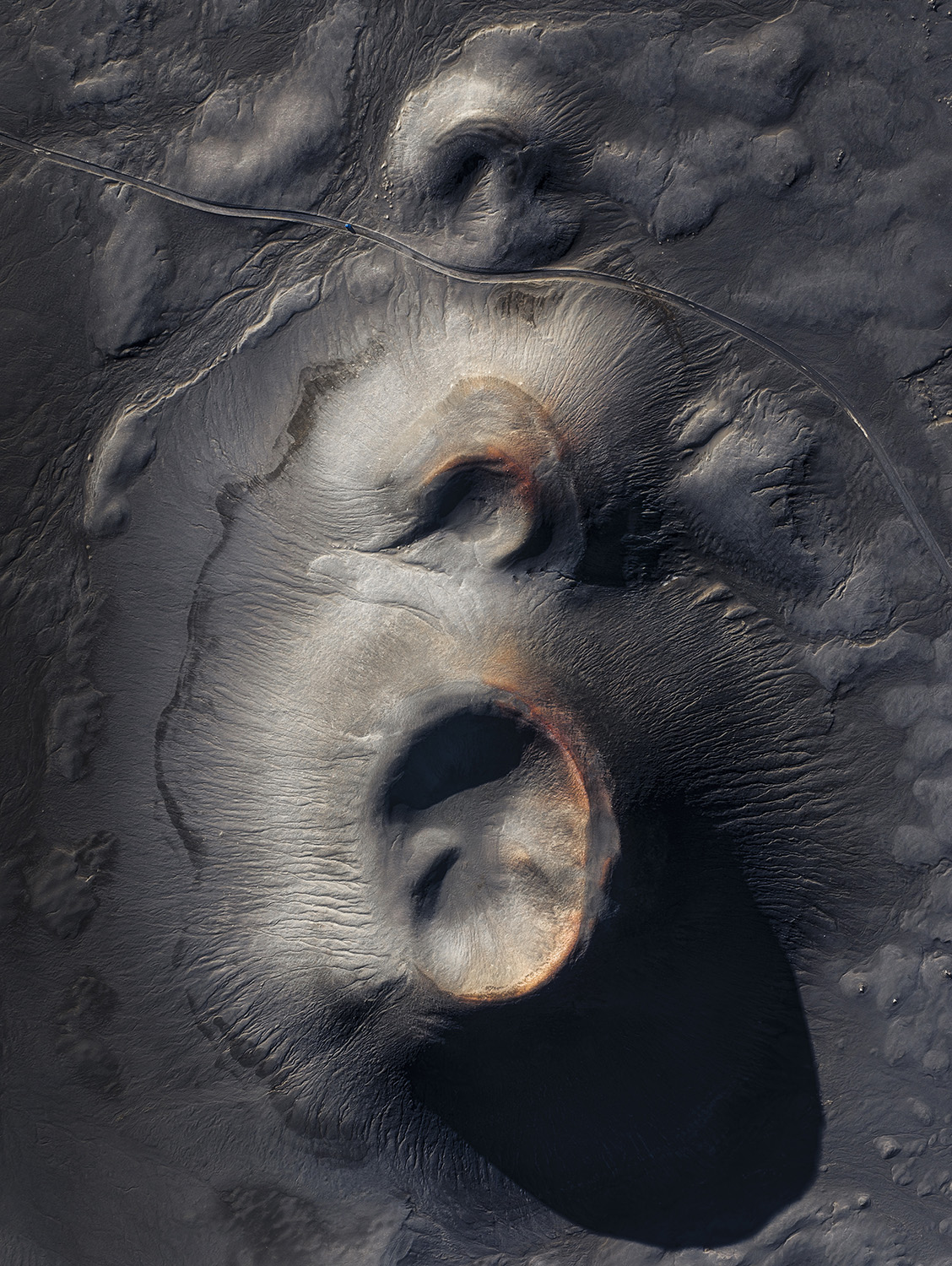 Aerial view of volcanic craters with rust-colored rims on dark ash-covered ground, with a road curving past.