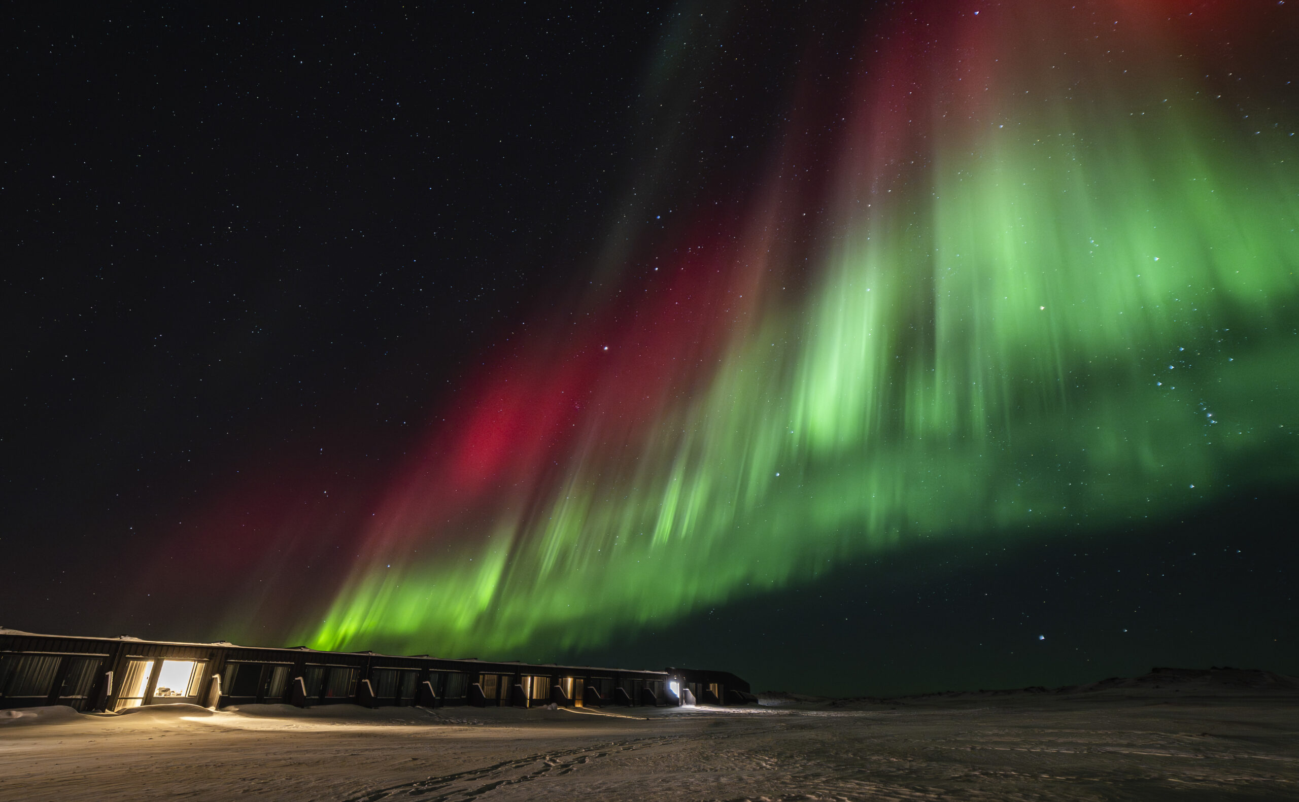 Aurora boreale verde e rossa sopra una pianura innevata, con una lunga costruzione bassa all'orizzonte.