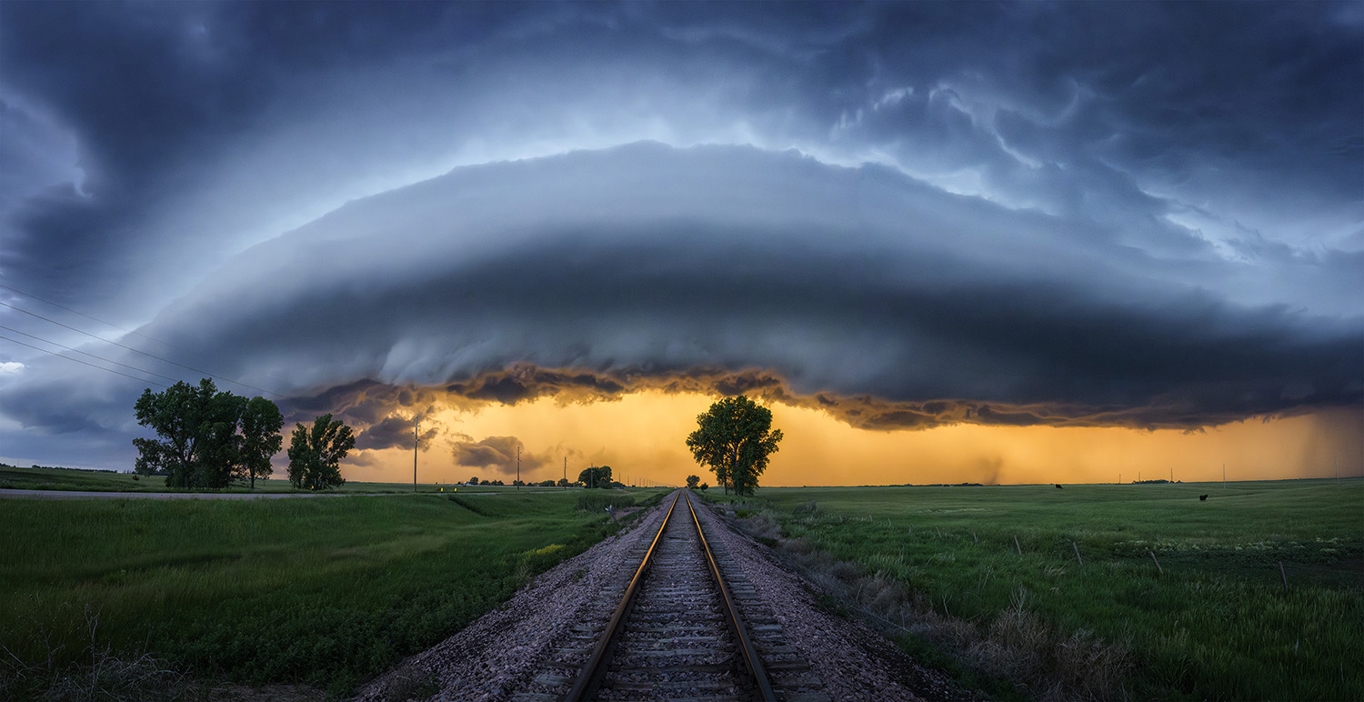 Shelf cloud arching over a railway track and green fields at sunset.
