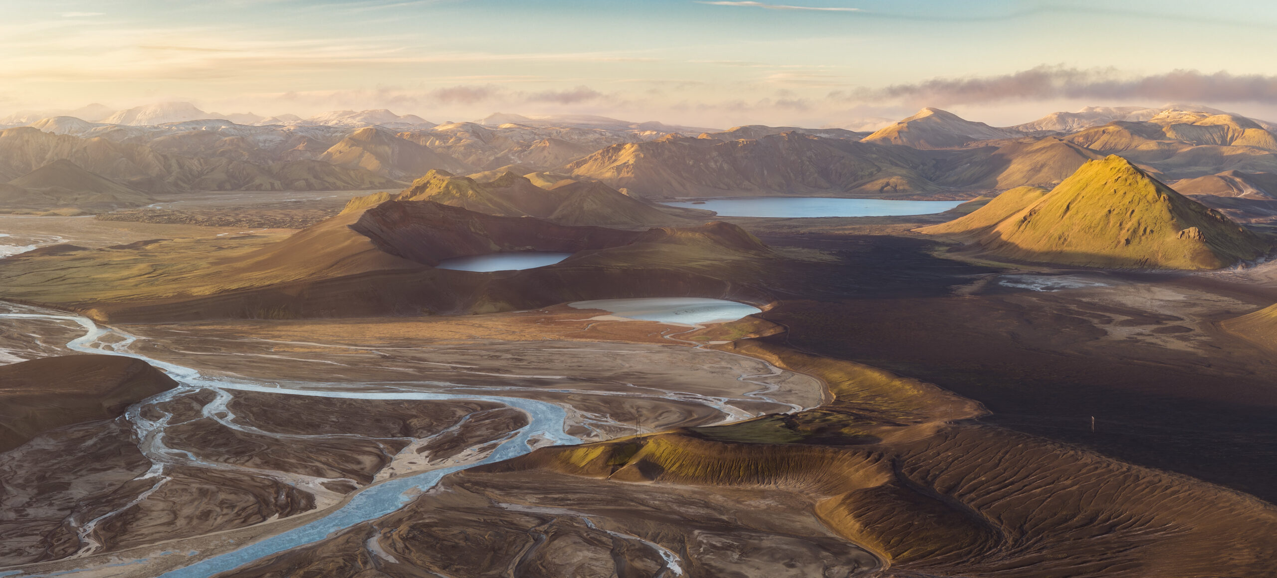 Veduta aerea degli altipiani islandesi con fiumi intrecciati, piccoli laghi e montagne dai colori caldi sotto un cielo chiaro.